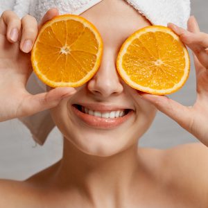 Mujer con dos rodajas de naranja sobre los ojos, representando el efecto antioxidante que tiene este cítrico, ya que la imagen acompaña un tratamiento cosmético antioxidante de un centro de estética